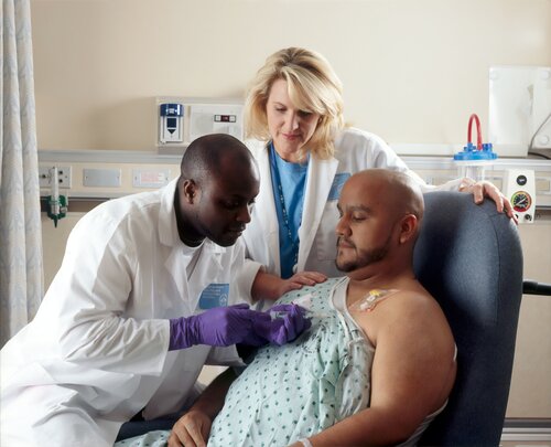 A Hispanic male patient receives Chemotherapy from a African-American Nurse through a port that is placed in his chest area. A caucasian female nurse looks on.